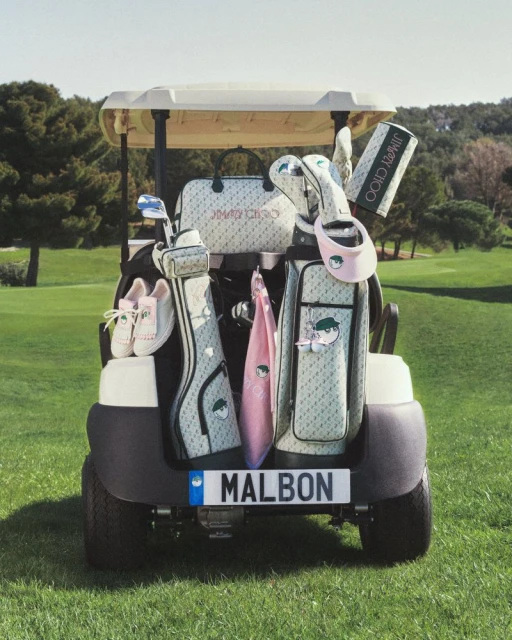 Golf cart filled with golf bags and accessories, featuring a Jimmy Choo handbag and Malbon license plate, set on a lush green golf course.