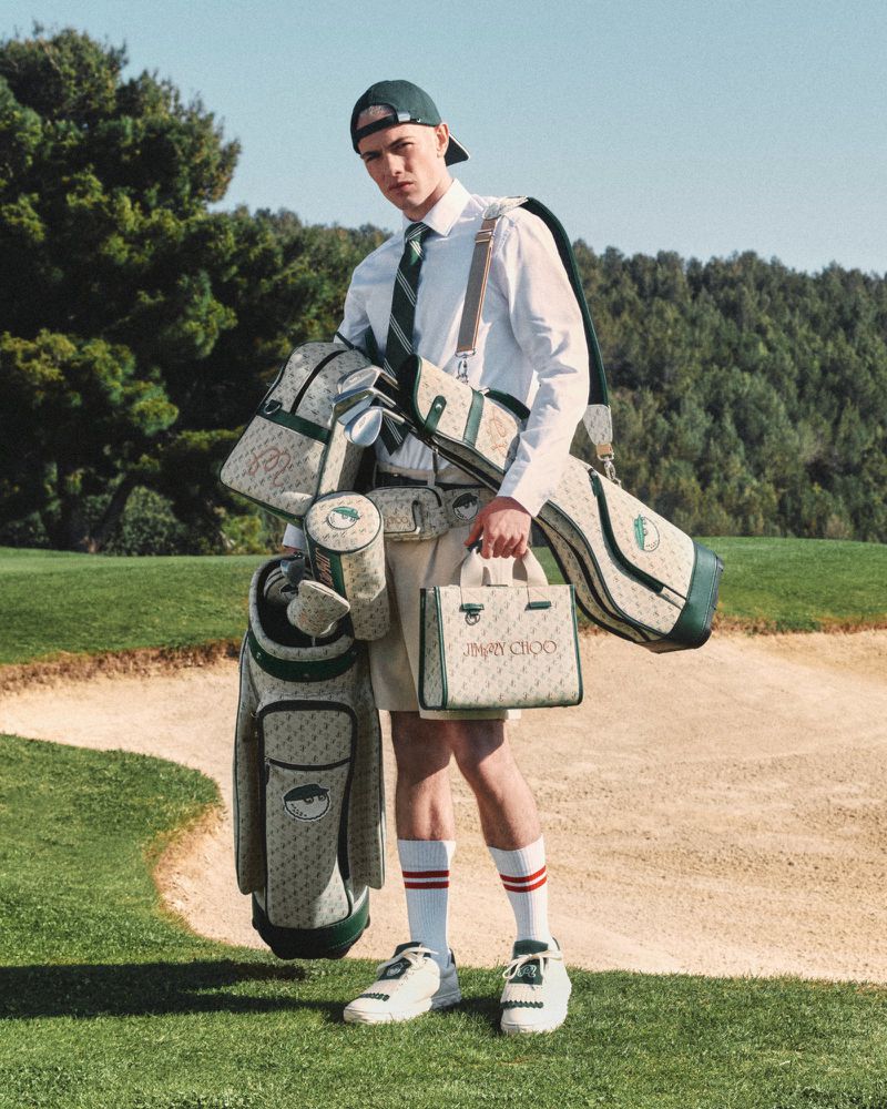 Male model wearing a green cap and shorts, carrying golf bags and accessories with the Jimmy Choo logo on a golf course.