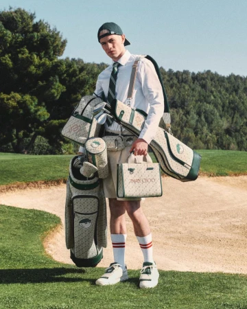 Male model wearing a green cap and shorts, carrying golf bags and accessories with the Jimmy Choo logo on a golf course.