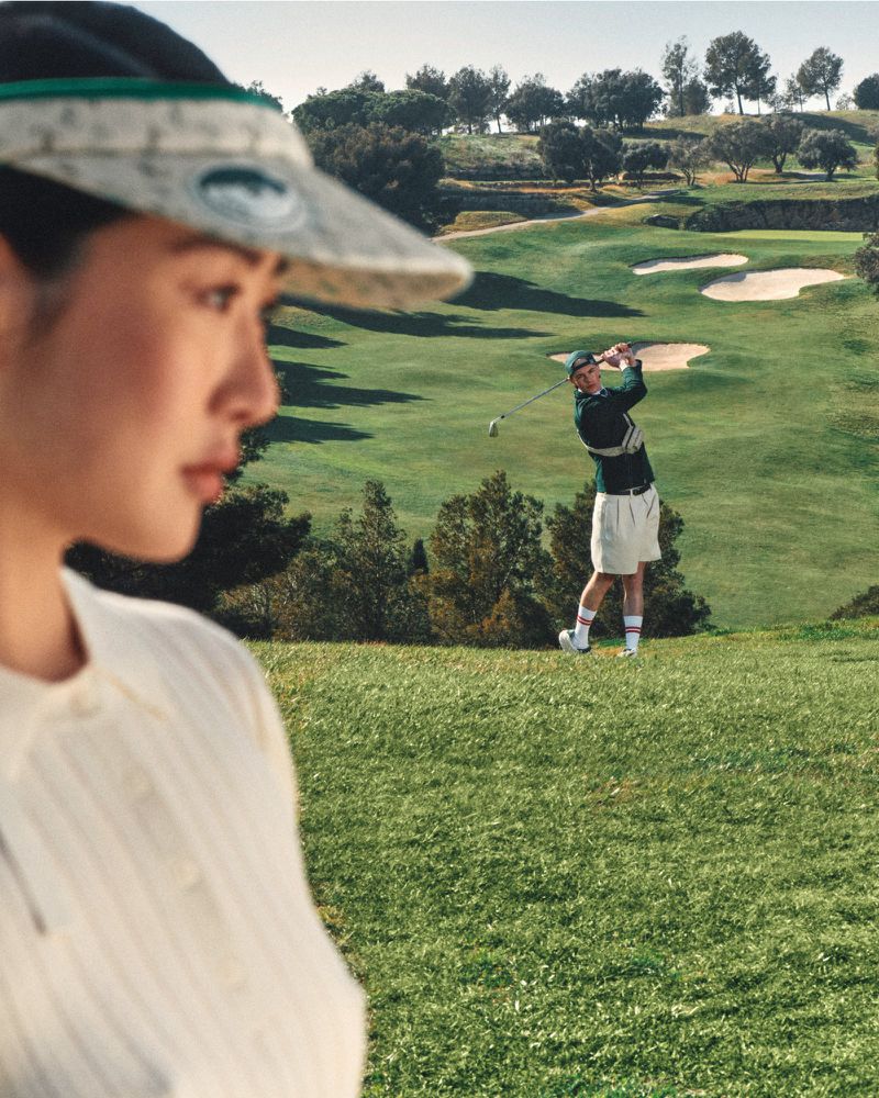 Woman in visor with golfer teeing off in background on green golf course with sand traps and trees on a sunny day, close-up.