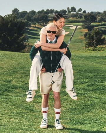 A man gives a woman a piggyback ride on a golf course; she holds a golf club. They are dressed in athletic clothing in a green landscape.