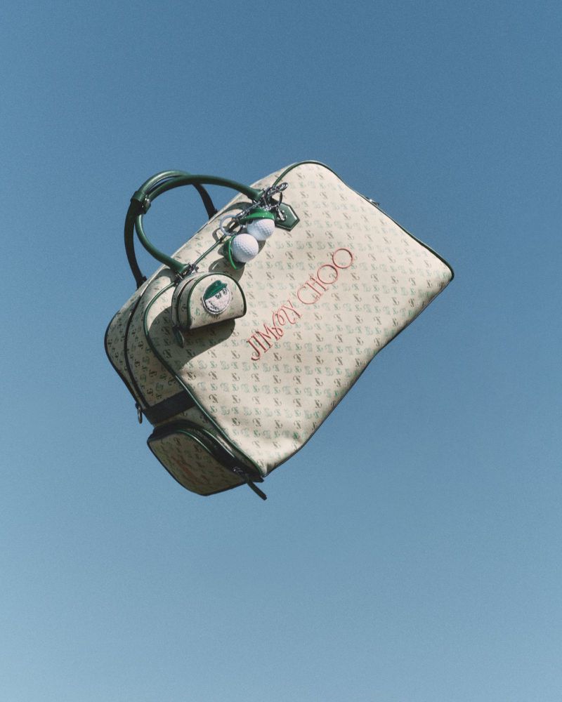 Tan patterned Jimmy Choo tote bag floats against a clear blue sky, featuring green trim, handles, and a decorative golf ball keychain.