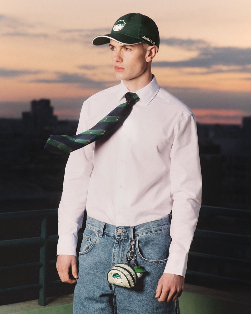 Adult male in a white button-down shirt, blue jeans, baseball cap, and tie stands outdoors with a cityscape at dusk.