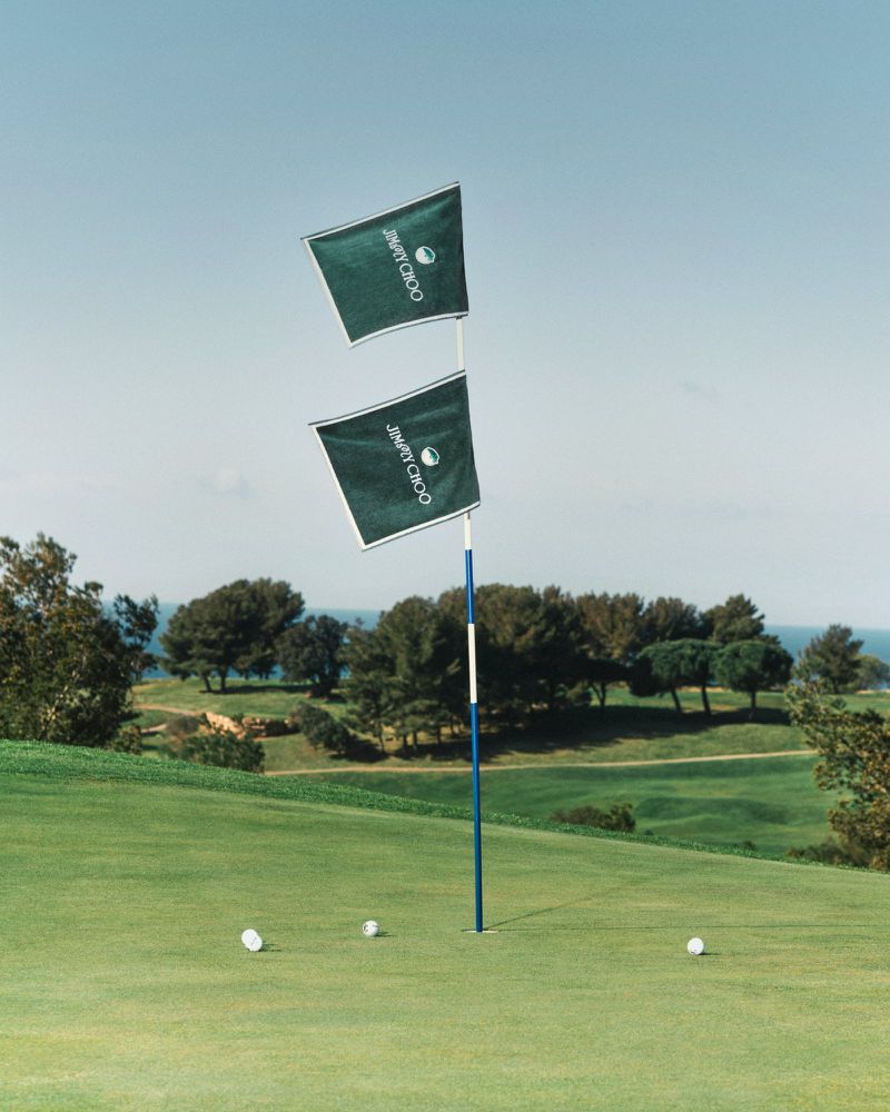 Golf course putting green with Jimmy Choo branded flags on the hole, surrounded by short green grass, trees, and a blue sky.