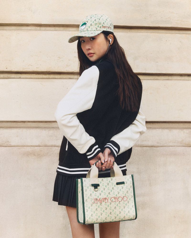 A fashionable young Asian woman stands against a textured wall wearing a baseball cap, varsity jacket, and carrying a Jimmy Choo handbag.