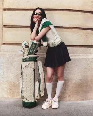 Smiling woman in sunglasses leaning on a golf bag wearing a white top, dark pleated skirt, and green sweater