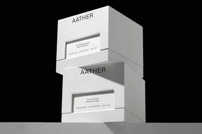 Two white AATHER candle boxes stacked against a black background, showing product names 'The Wanderer Resurr' and 'As the Sue Sets'.