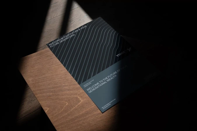 Close-up of marketing brochure on wooden surface with diagonal sunlight. Brochure features a modern design with geometric line pattern.