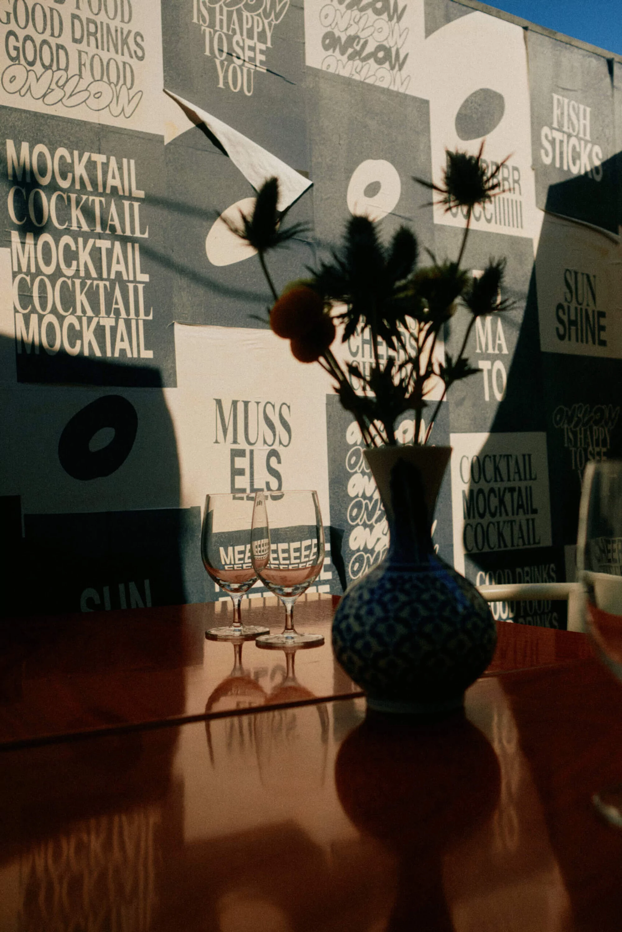 Still life of wine glasses, vase with thistles, and an orange fruit set on a red table, with a graffiti wall as a backdrop.