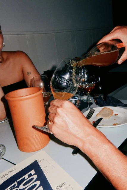 Person pouring amber-colored liquid into a wine glass at a table with a drink menu, a brown container, silverware, and other wine glasses.