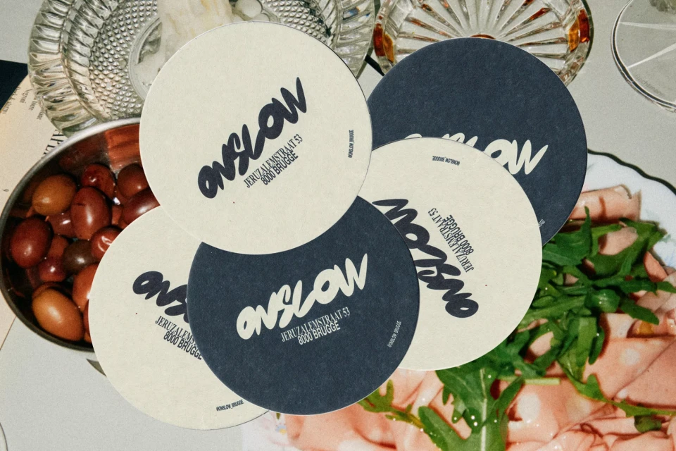 Arrangement of Onslow restaurant coasters, bowl of olives, cured meats, and glass with liquor on a white surface.