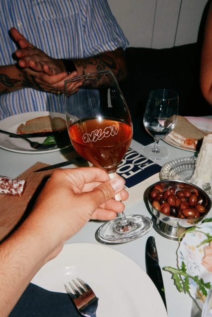 Eye-level shot of a wine glass with liquid inside, held at a dining table with food, plates, and silverware, creating a convivial atmosphere.