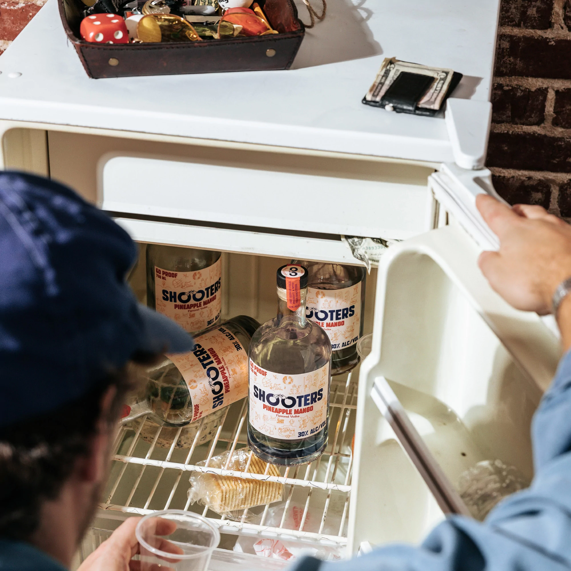 Messy refrigerator interior with liquor bottles, an opened case of crackers, a blue-capped person, and a leather container on top.