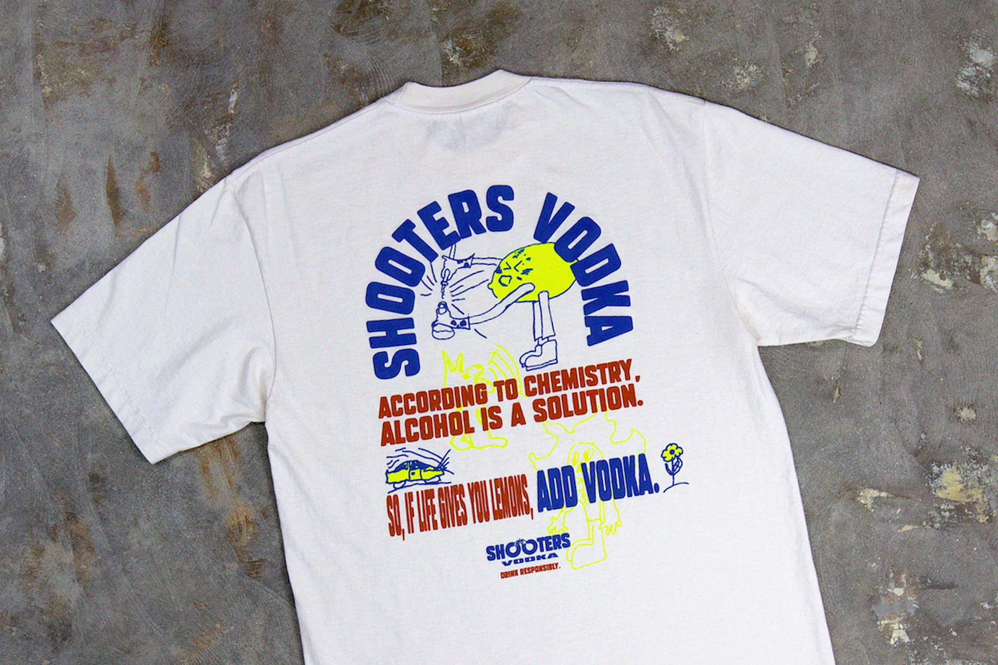 A white t-shirt laid flat showcasing a blue and yellow cartoon design promoting Shooters Vodka with humorous chemistry-themed text.