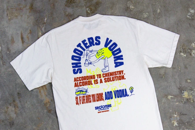 A white t-shirt laid flat showcasing a blue and yellow cartoon design promoting Shooters Vodka with humorous chemistry-themed text.