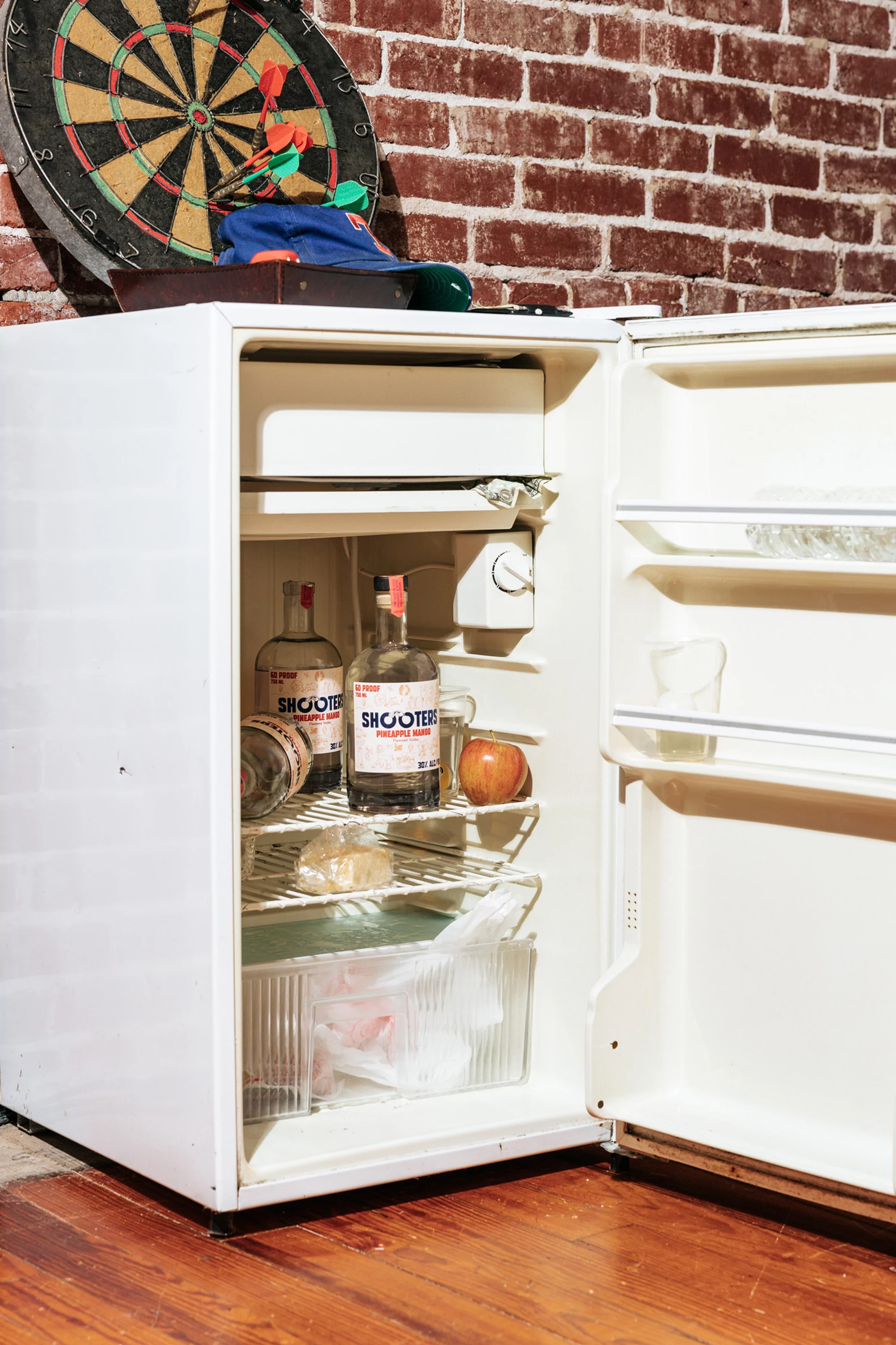 Opened white mini refrigerator containing alcohol bottles, an apple, and other objects, sits against a red brick wall with a dartboard.