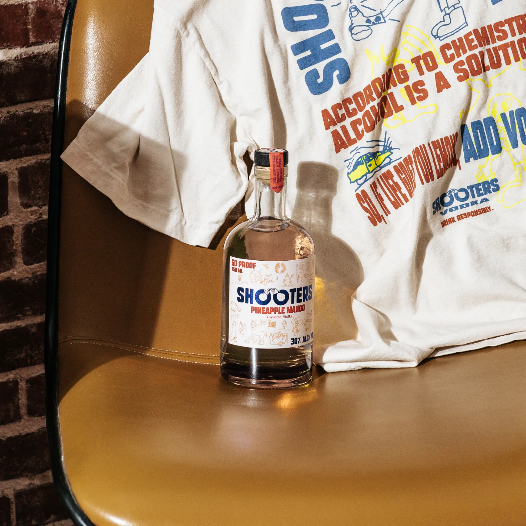 Shooters Vodka Pineapple Mango bottle on a brown chair with a graphic t-shirt, capturing a modern, lifestyle product shot.