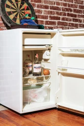 An open white mini fridge containing Shooters Vodka bottles, an apple, and packaged items against a red brick wall
