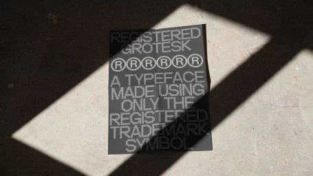 A black poster featuring pixelated typeface designs lying flat on a textured concrete surface under diagonal shadows.