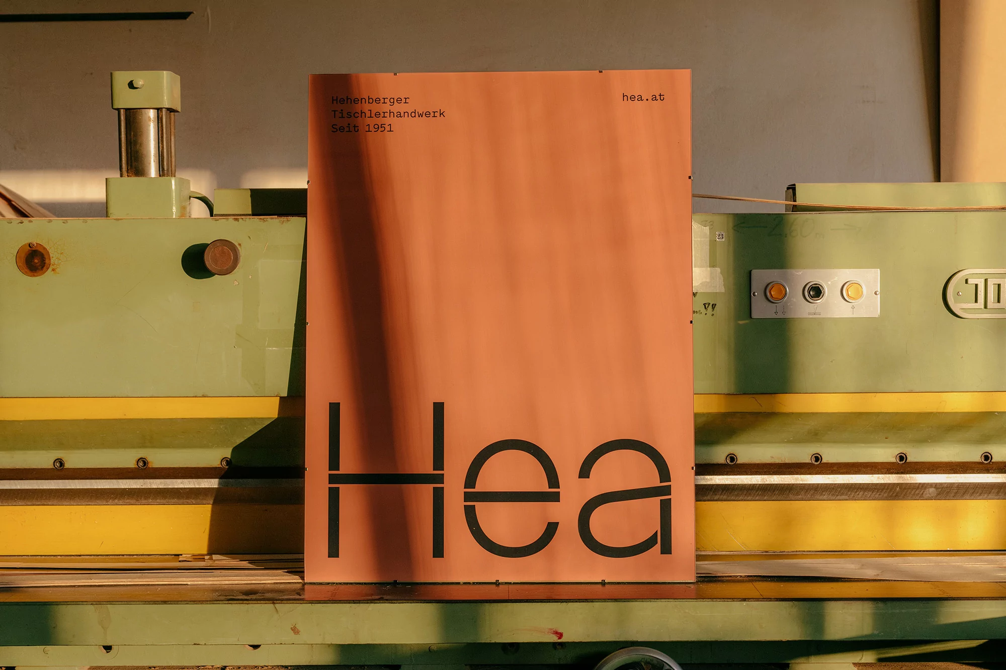 Close-up of an orange poster with black text 'Hea', against a green and yellow industrial machine in a workshop or factory setting.