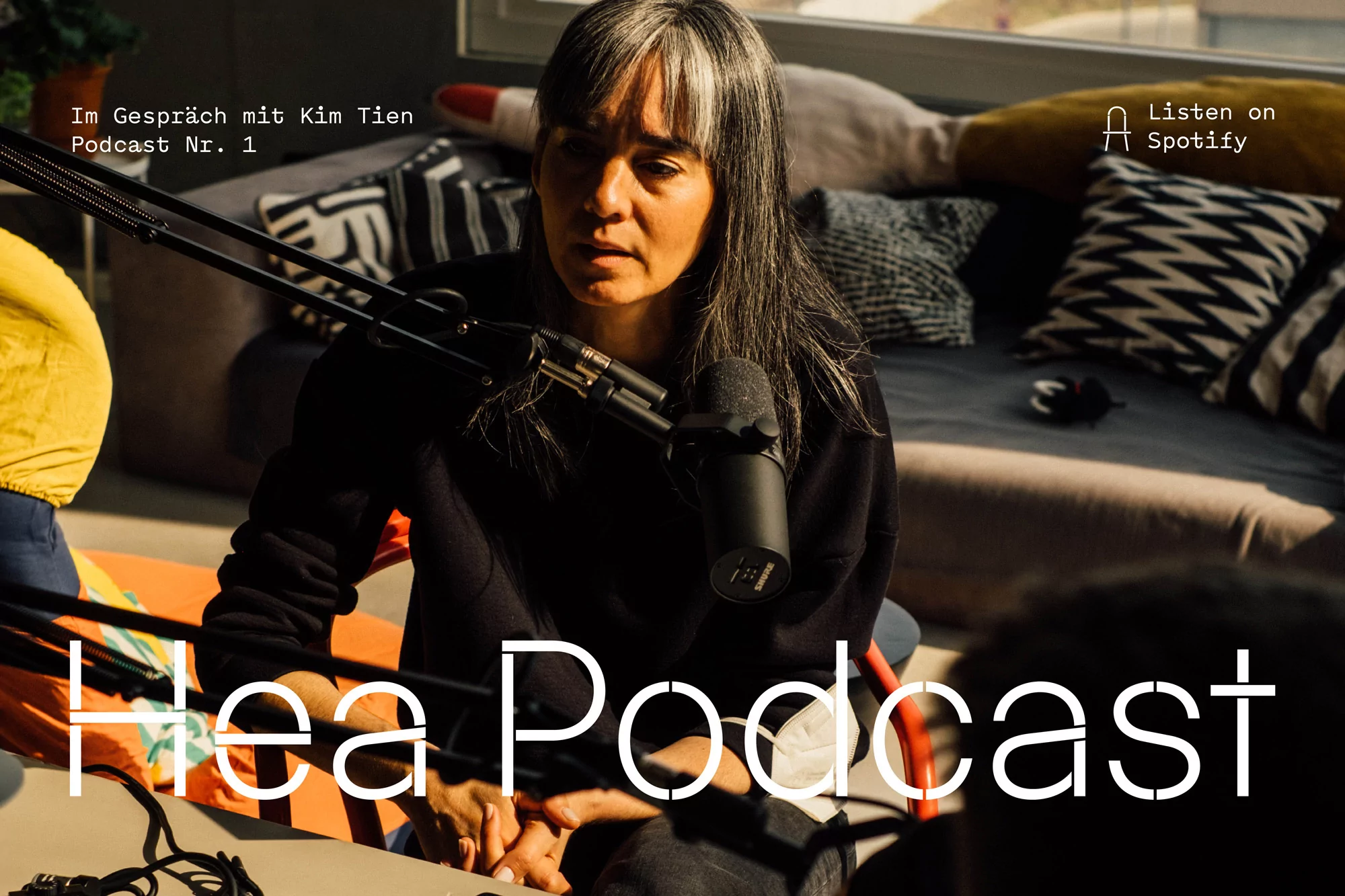 Woman records podcast in living room with professional microphone, decorative pillows, natural light, casual clothing, and streaming text overlay.