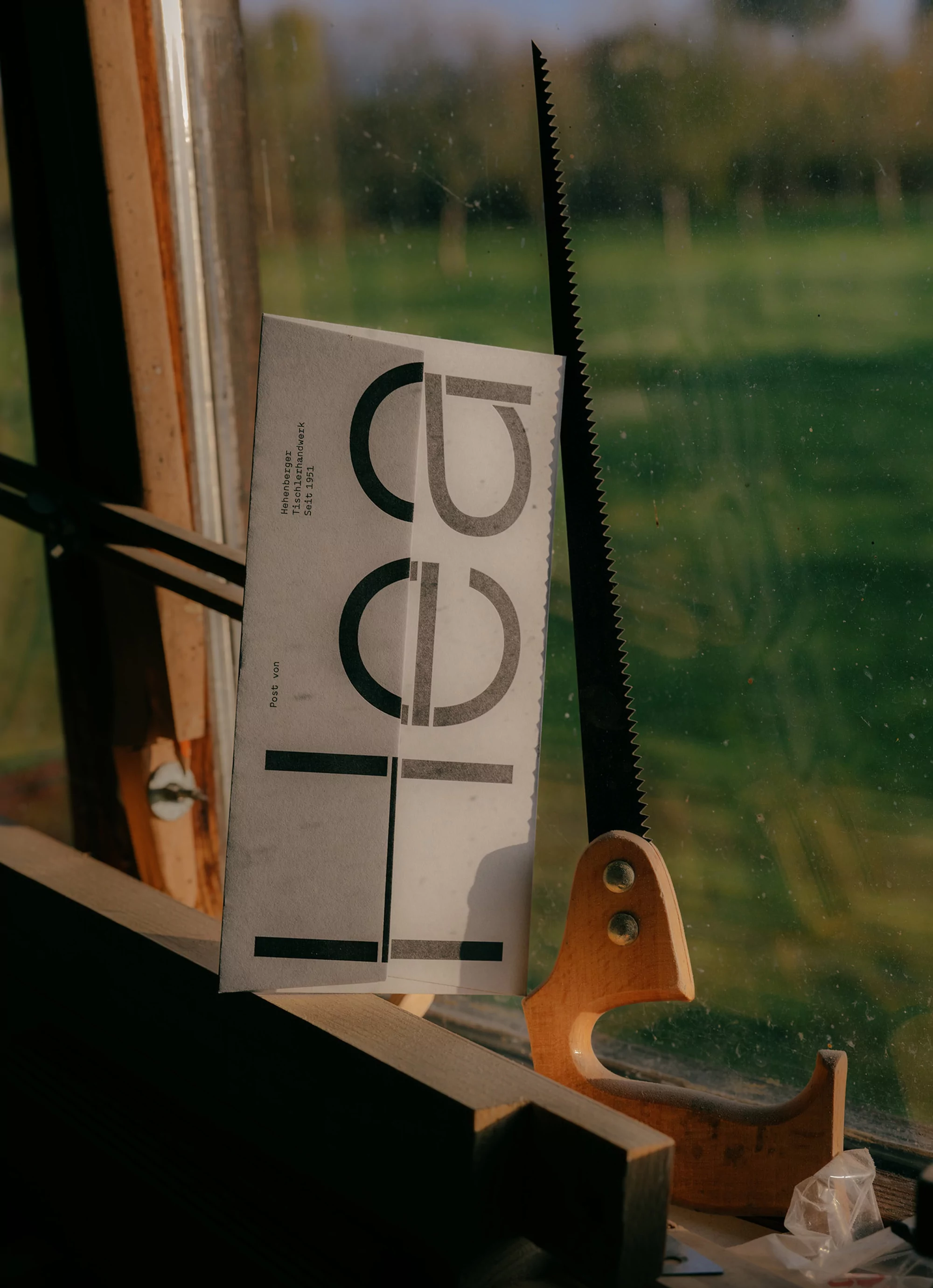 Still life photograph capturing a book, hand saw, and carpentry elements in a naturally lit, cluttered environment. Wood and printed text visible.