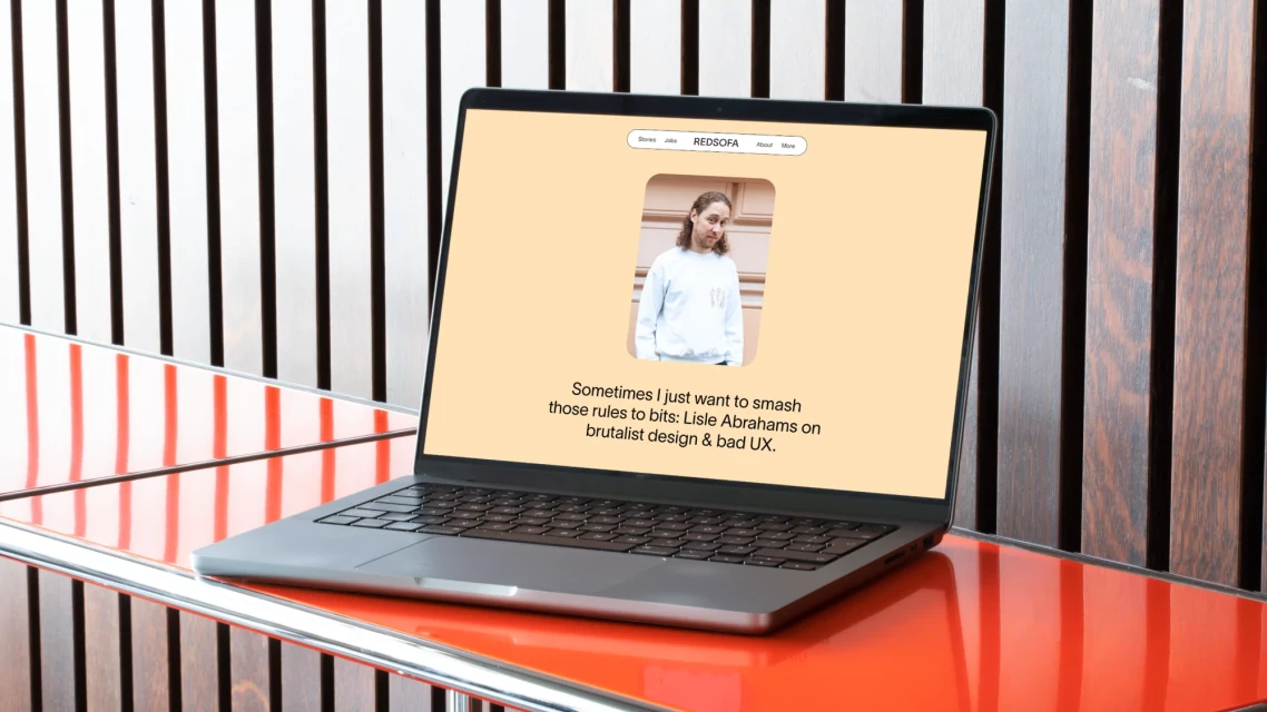 Laptop displaying a website featuring a photo of a person and text about design on a table with wooden slats, indoors during daytime.