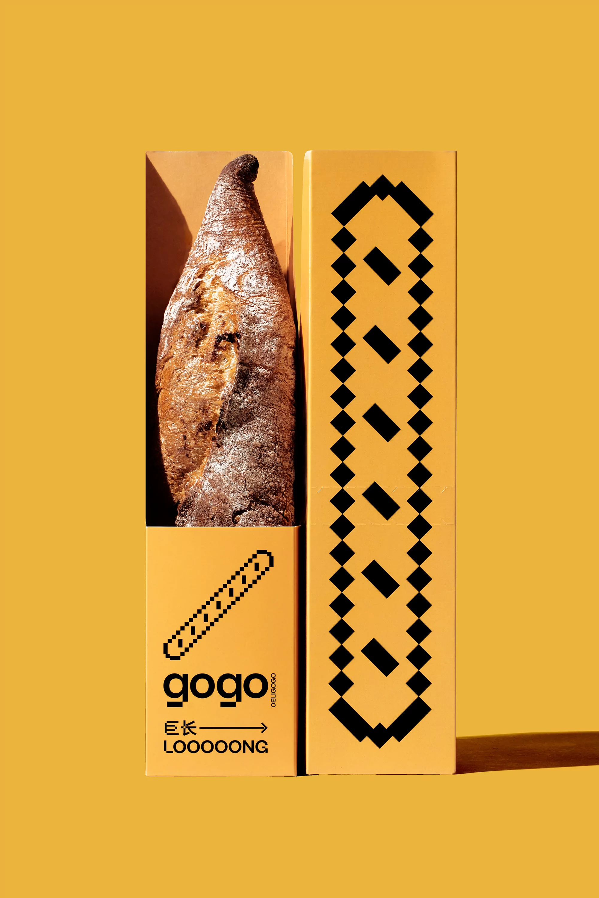 A brown baguette rests inside an opened tan cardboard box with a black patterned design against a mustard-yellow background in a studio shot.