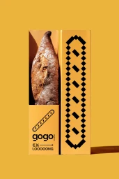 A long artisanal loaf of bread in a yellow branded Gogo box standing against a matching yellow background