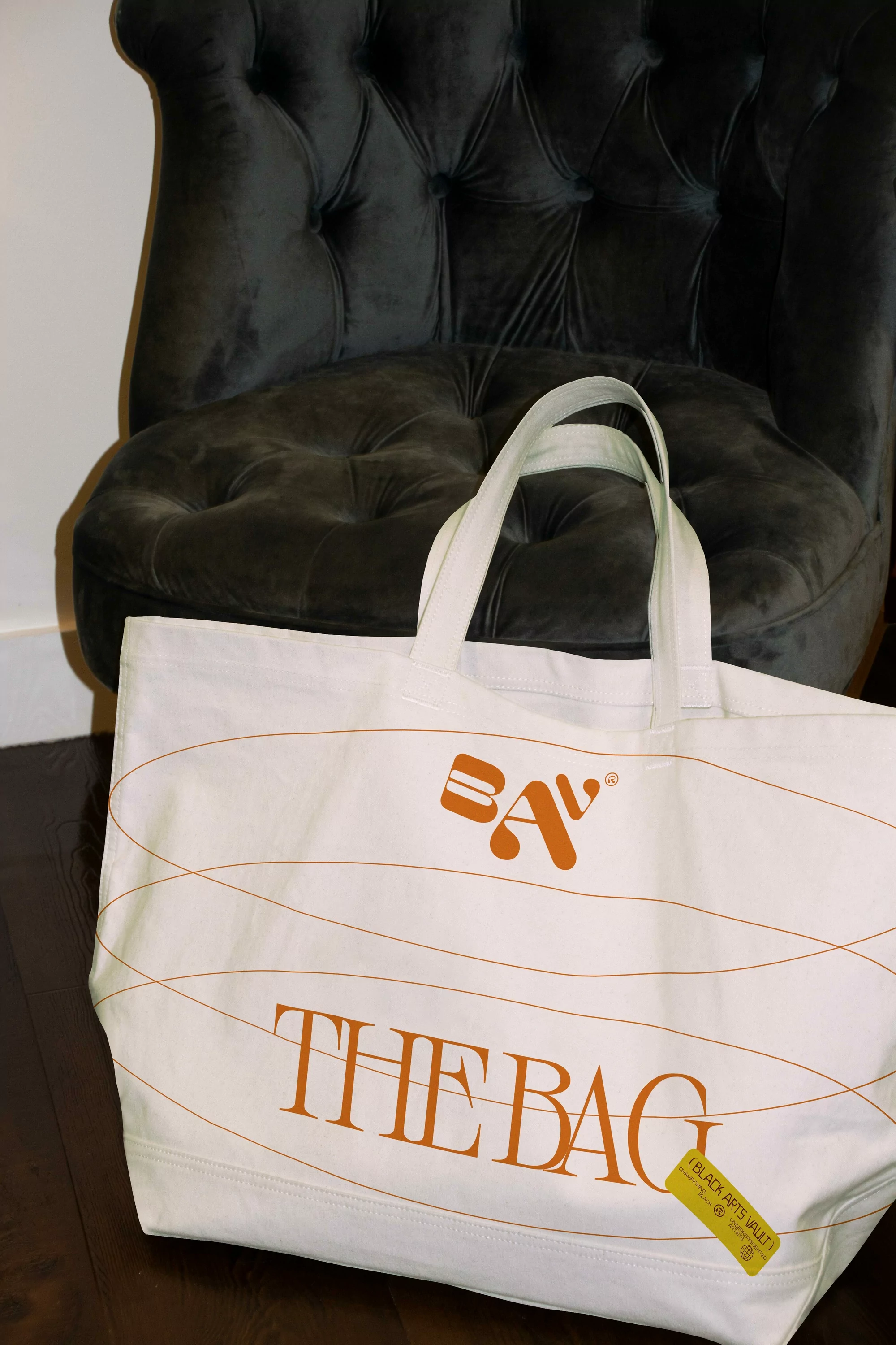 White canvas tote bag with brown decorative lines and "The Bag" text, placed in front of a dark grey tufted chair on a dark wood floor.