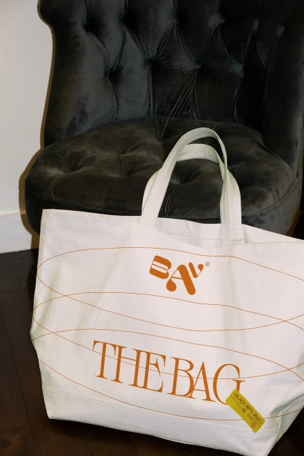 White canvas tote bag with brown decorative lines and "The Bag" text, placed in front of a dark grey tufted chair on a dark wood floor.