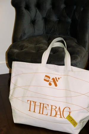 White canvas tote bag with brown decorative lines and "The Bag" text, placed in front of a dark grey tufted chair on a dark wood floor.