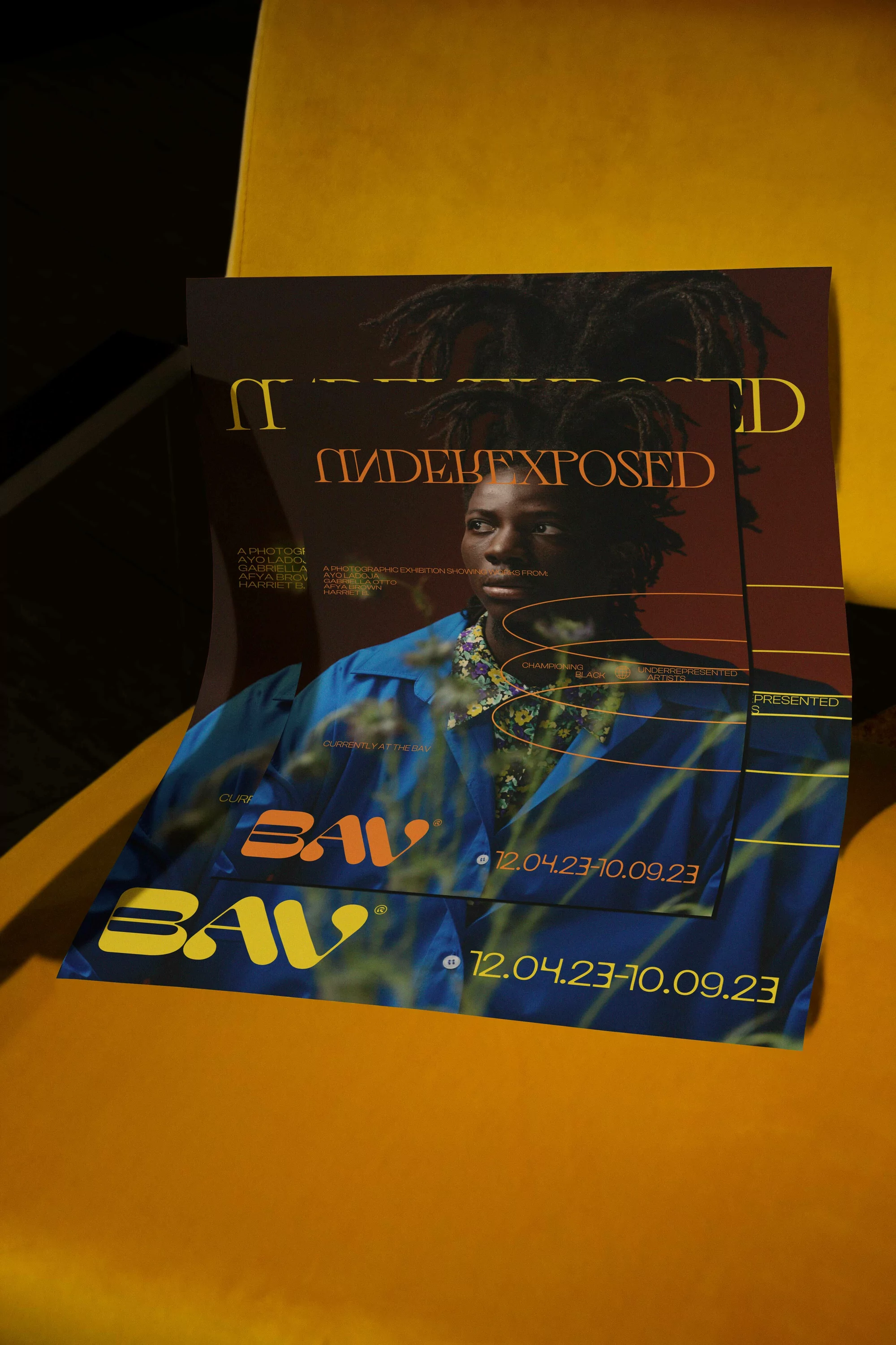 Promotional posters of a man with dreadlocks, wearing a blue shirt and gold jewelry, displayed on a yellow chair in a studio setting.