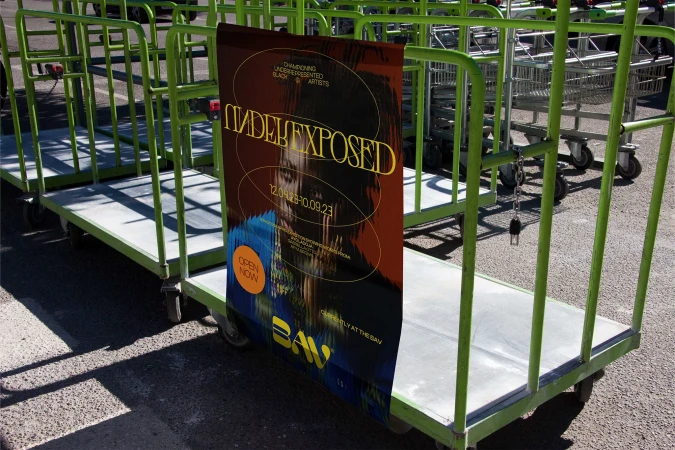 Poster on a green metal cart advertises 'Under Exposed' art exhibition at BAY. The poster has abstract art and text.