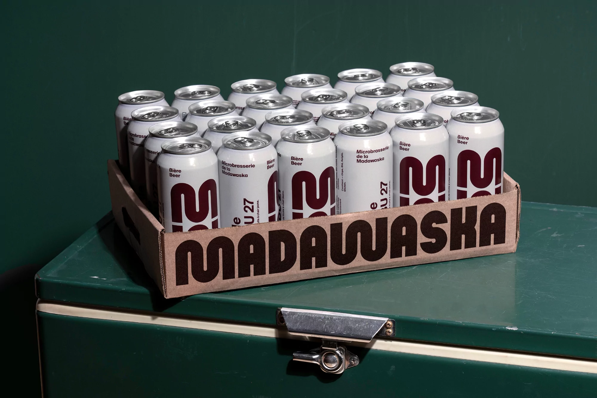 Close-up of a cardboard box filled with cans of Madawaska Bière Beer on a green cooler. Dark green background.