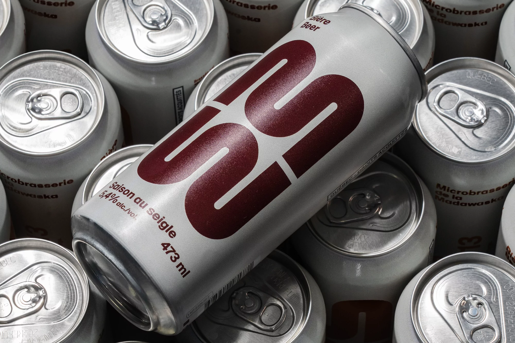 Multiple silver aluminum cans of beer are stacked in a close-up, high-angle shot. The main can features burgundy text. Product photography.