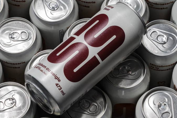 Multiple silver aluminum cans of beer are stacked in a close-up, high-angle shot. The main can features burgundy text. Product photography.