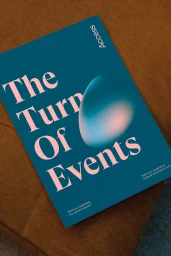 A blue book cover with pink serif typography reading The Turn Of Events resting on a brown textile surface.