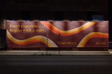 Large exterior mural with warm red and yellow wave graphics and typography reading Every experience begins with a story on a brick wall at night