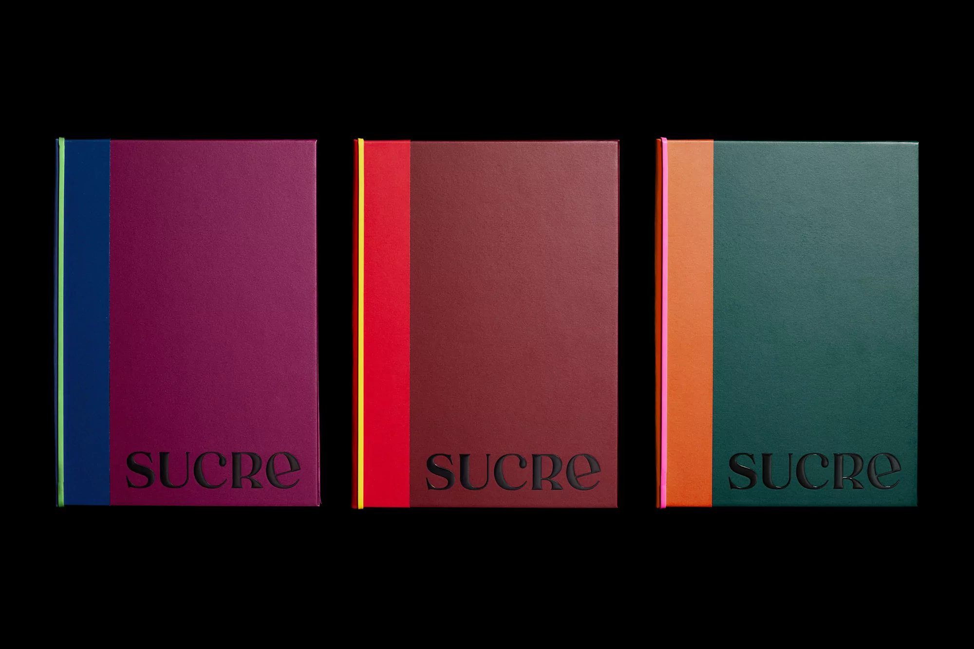 Three colorful notebooks sit side-by-side against a black backdrop, each featuring a unique color combination and the word 'SUCRE' embossed.