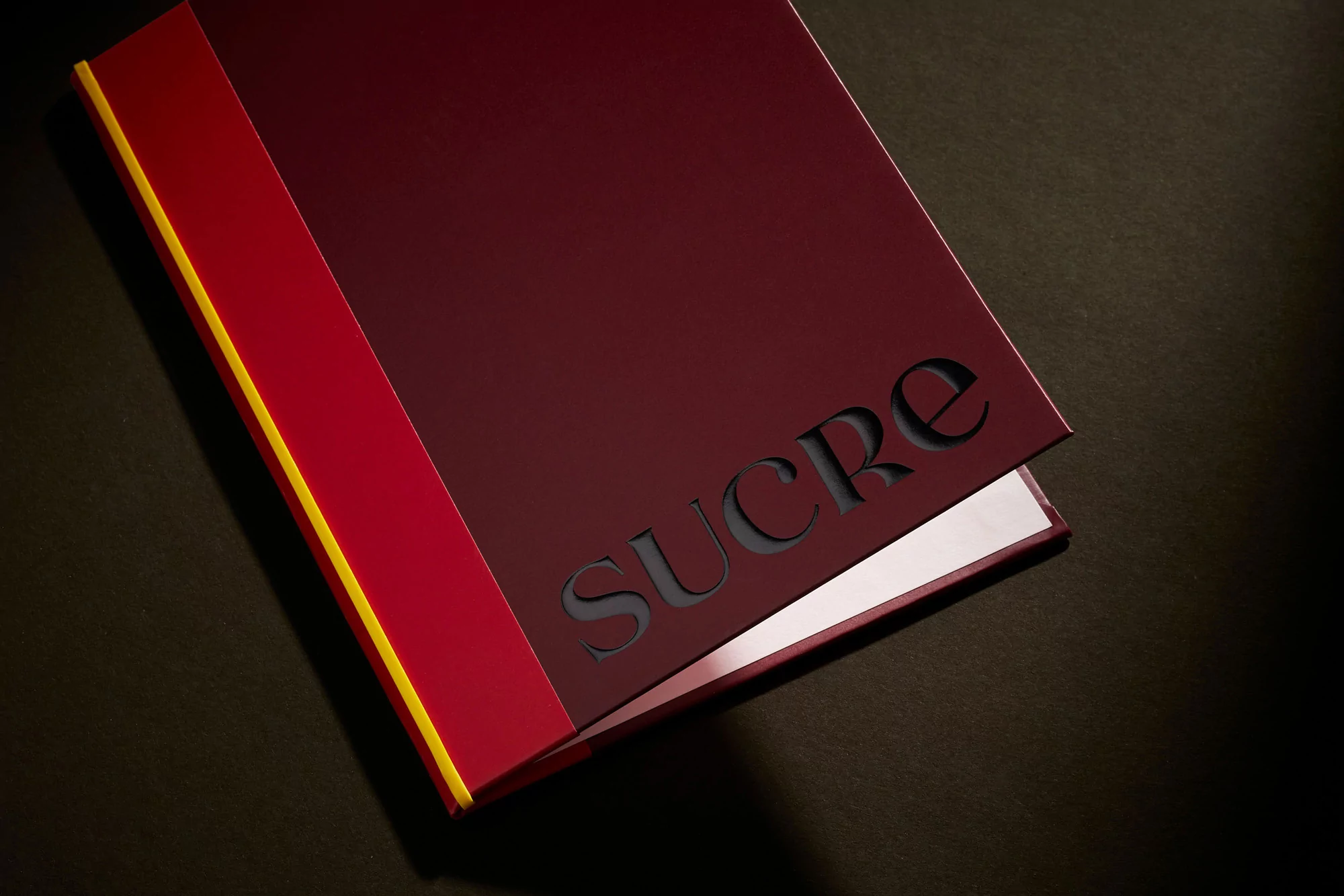 Burgundy-colored book with a red and yellow spine, featuring the word 'SUCRE' cut out from the cover, displayed on a dark surface.