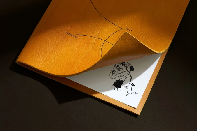 Yellow leather folder with black line art of Argentina and Italy, plus a person walking a dog illustration on white paper.