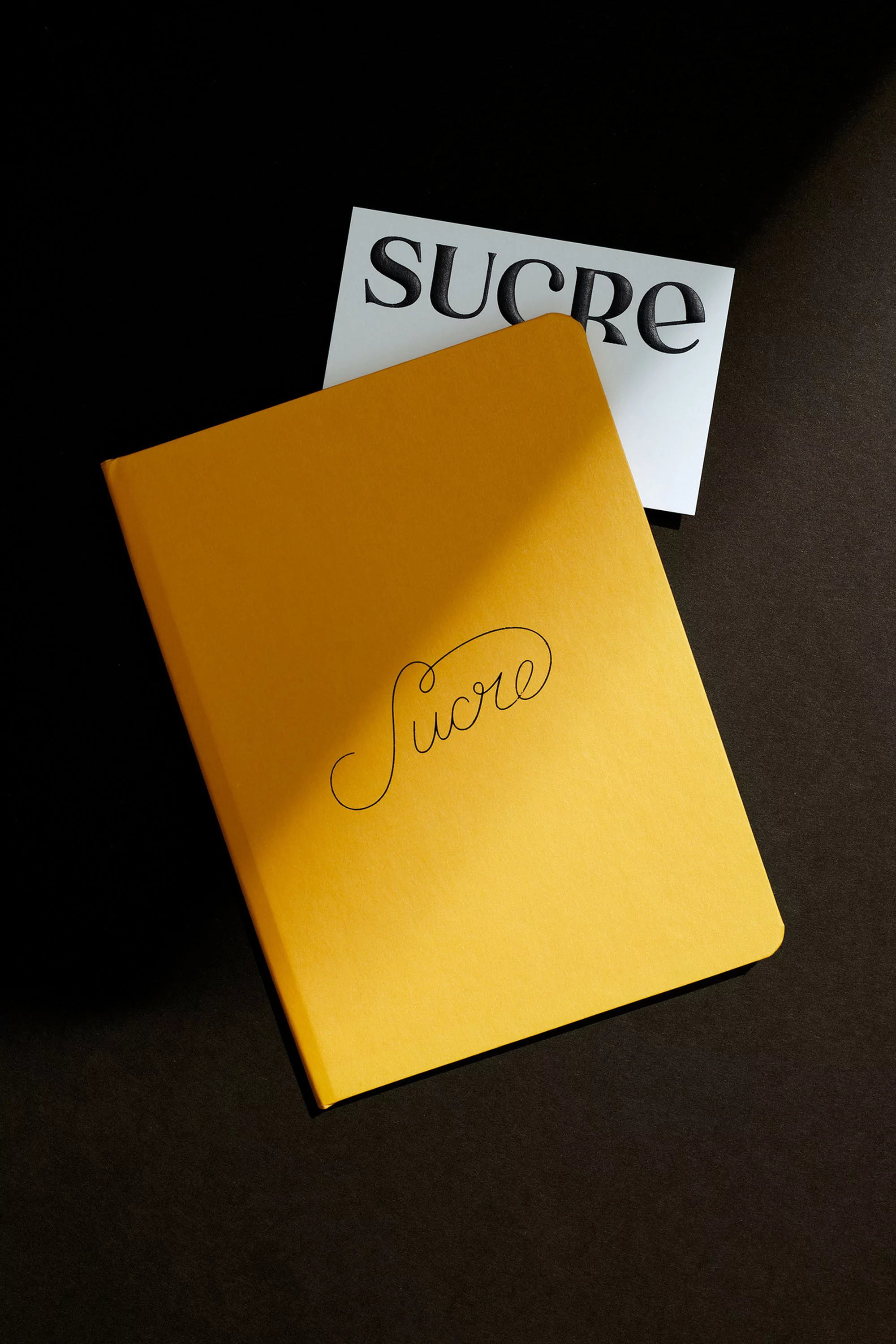 Yellow journal with 'Sucre' writing on top and a white card with the same word over a dark surface illuminated by a light.