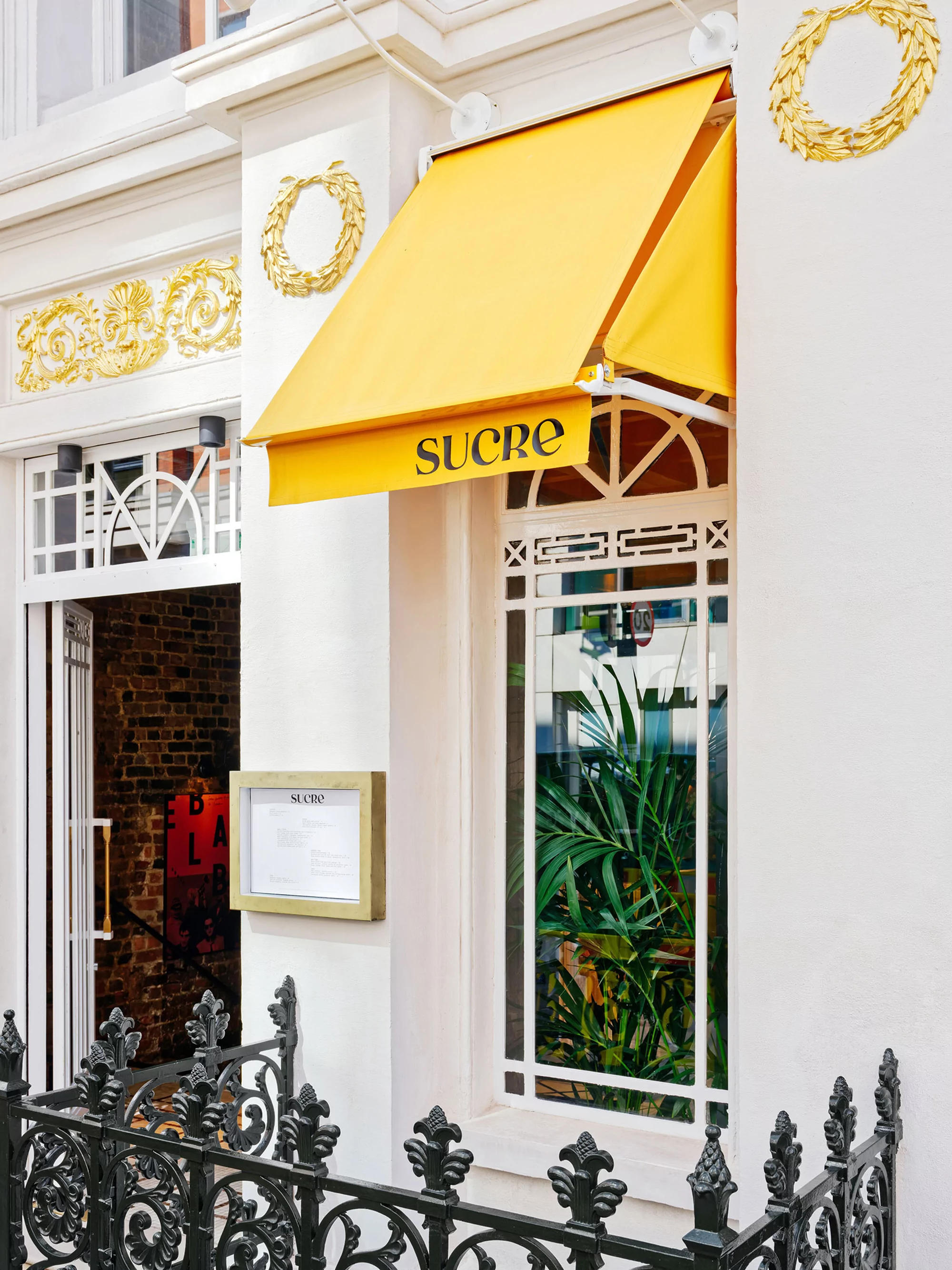 Exterior of the Sucre restaurant with a yellow awning and decorative gold wreaths on the building's facade.  Window shows plants inside.