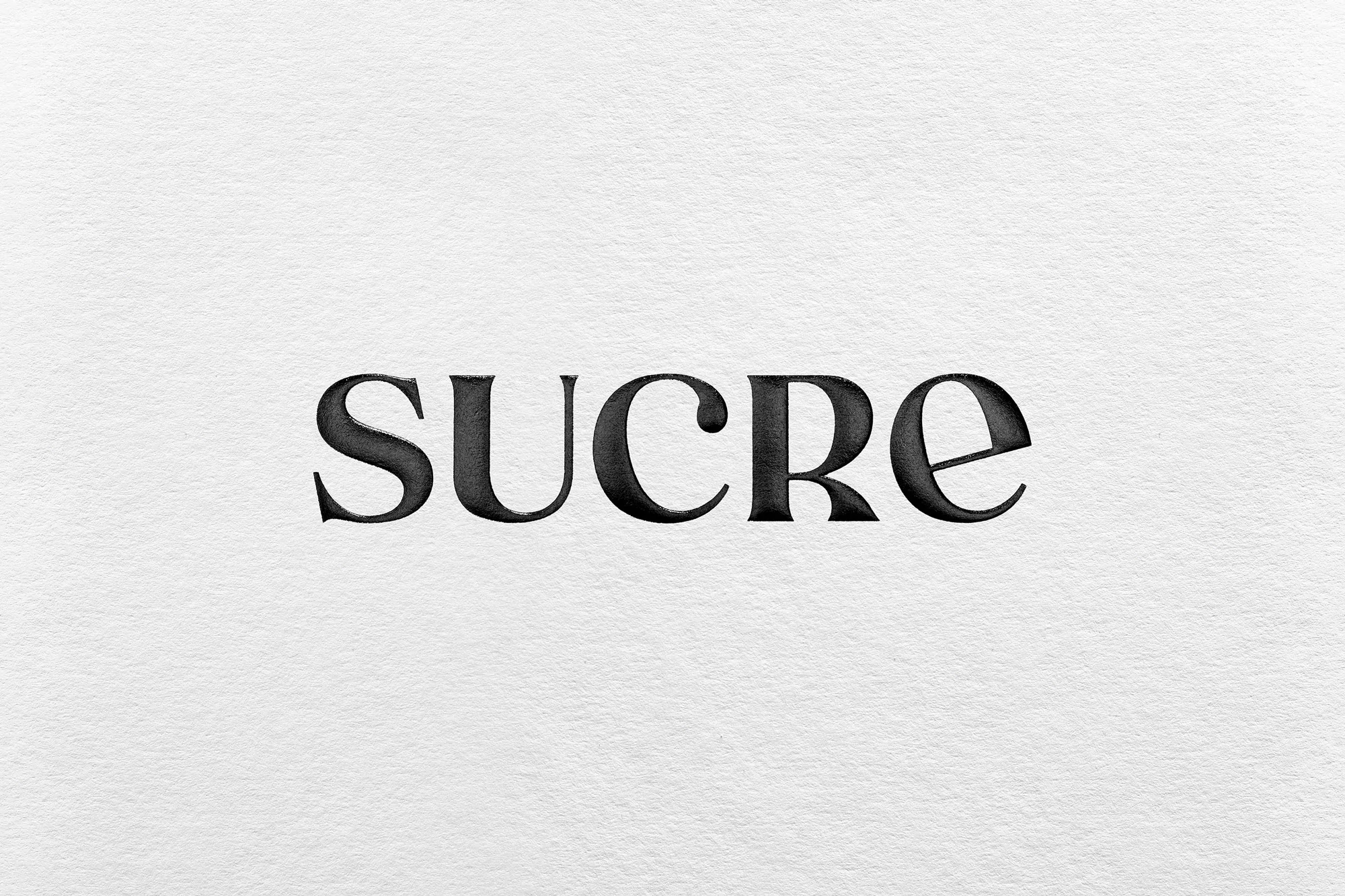 Elegant typography on textured paper. Close-up studio shot of the word 'SUCRE' in a serif typeface, displaying craftsmanship and simplicity.