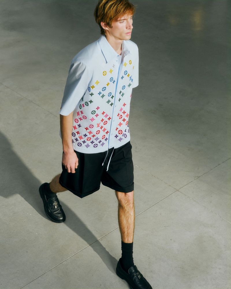 Man strides in a light blue button-down shirt with multicolored monogram pattern, black shorts, black socks and loafers against grey backdrop.