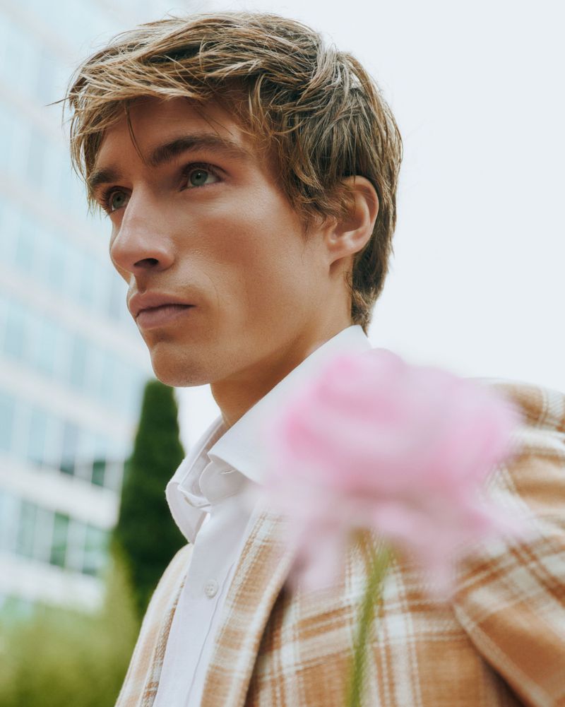 Portrait of a blond male model in a plaid blazer, posed outdoors with a flower in the foreground, set against a building backdrop.