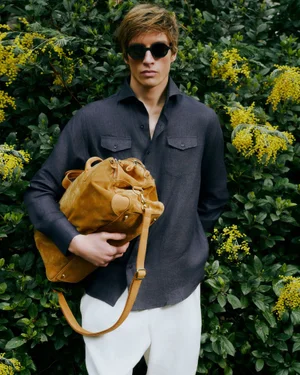 Male model wearing sunglasses and a dark button-down shirt holds a tan suede duffel bag before yellow flowering foliage.