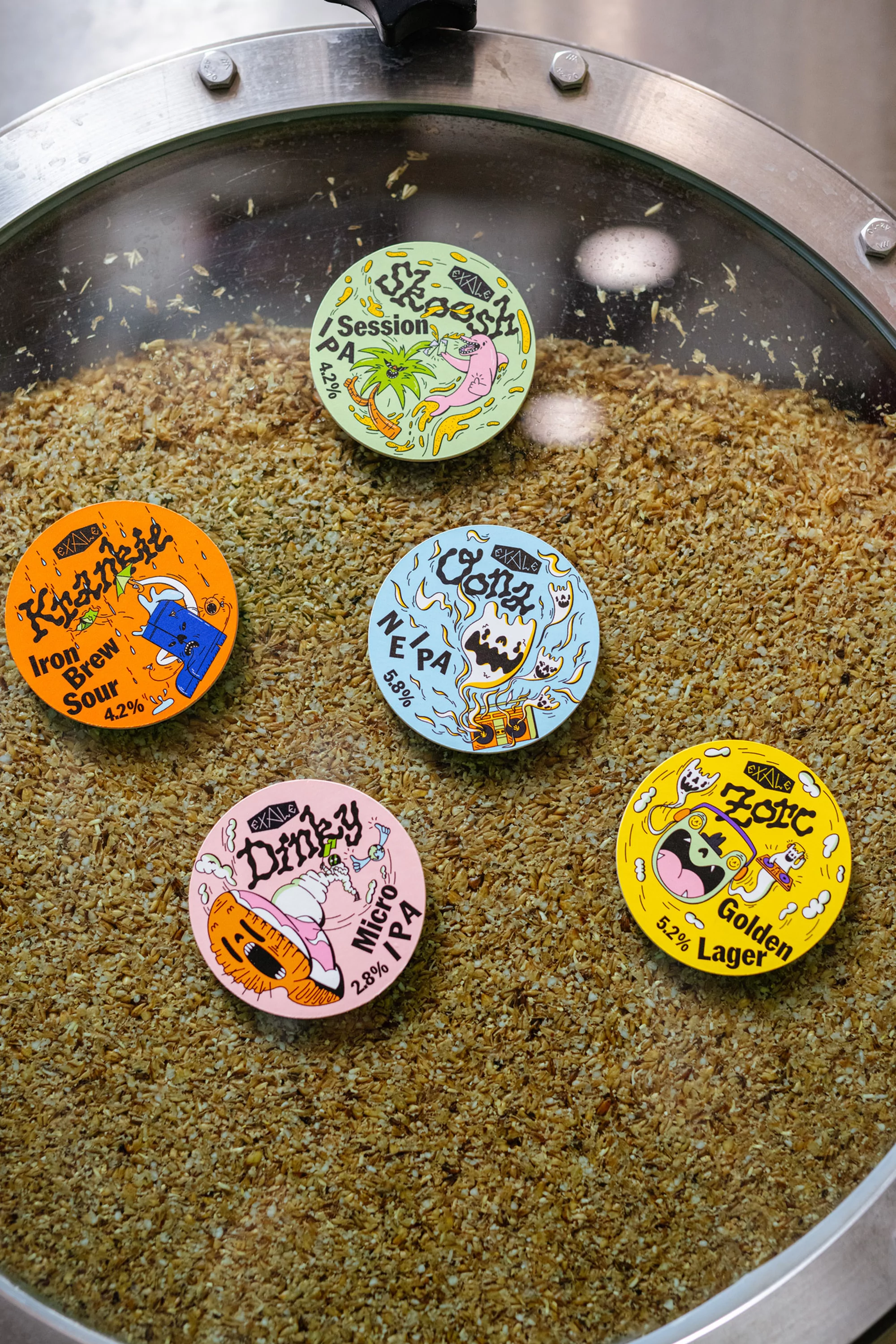 Close-up of five colorful beer labels in a brewing vat, featuring cartoon graphics and beer names like Skoosh, Krankie, and Zorc.