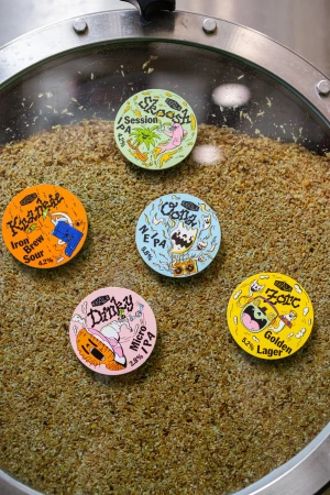 Close-up of five colorful beer labels in a brewing vat, featuring cartoon graphics and beer names like Skoosh, Krankie, and Zorc.